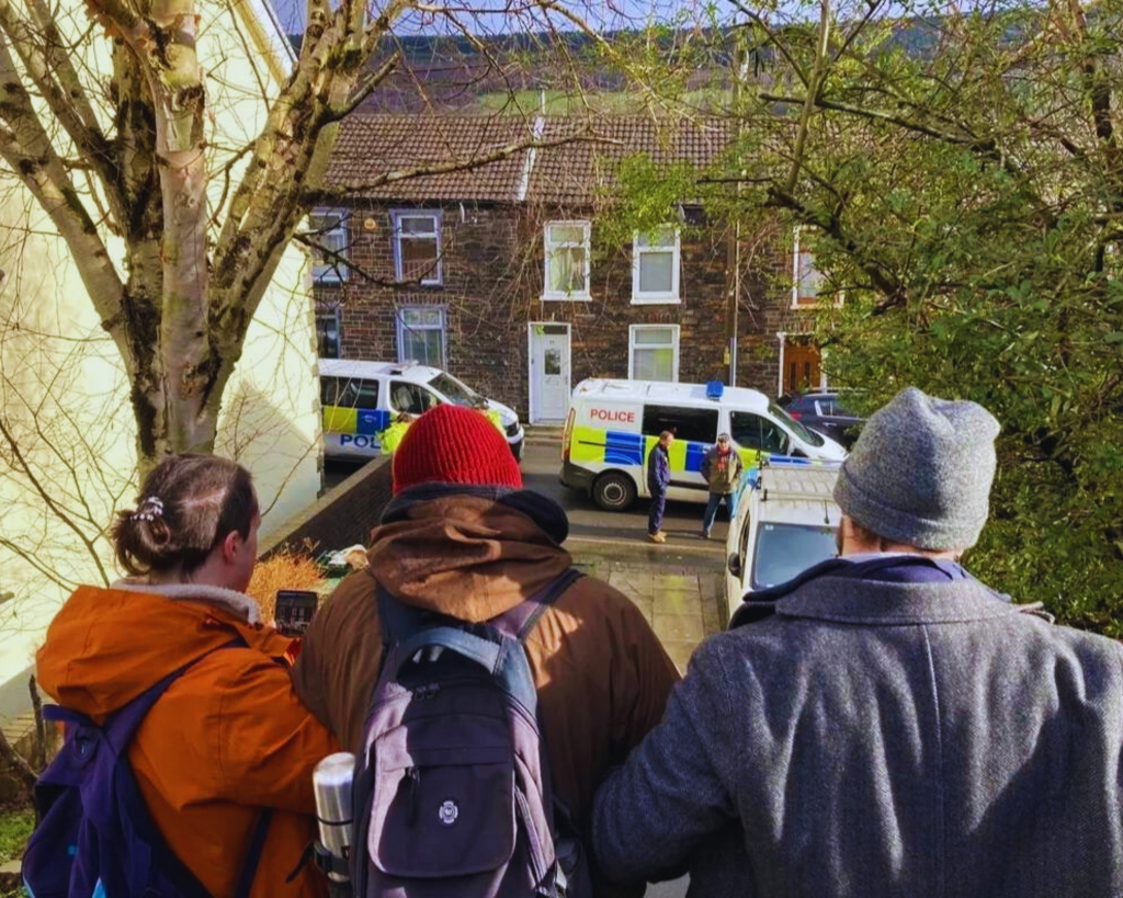 Photograph of three people, arms joined, facing two police vans on a quiet street