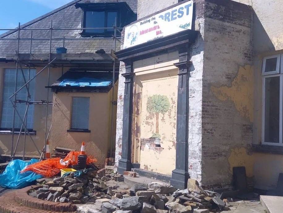 Photograph of a derelict looking building, the windows are covers, the main door is boarded over and there are piles of rubble and rubbish on the floor in front. The sign over the door is peeling off.