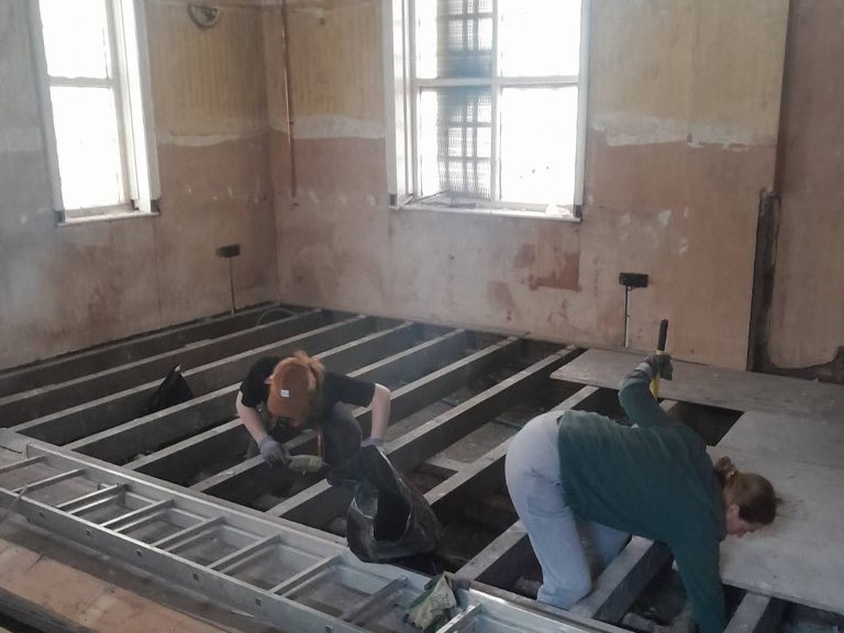 a photograph inside the stute, the walls are bare plaster, there are no floorboards and two comrades are cleaning debris from the floor