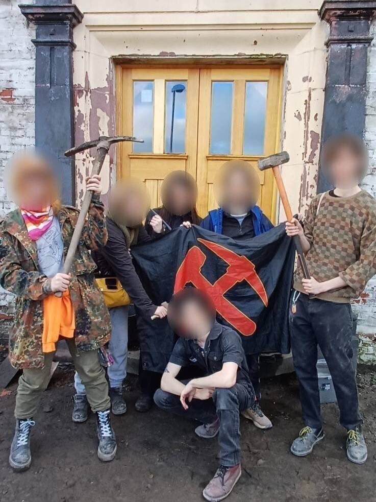 Photograph of 6 comrades grouped around a WUN flag in front of the miners' institute doors. They hare raising a pickaxe and sledgehammer in the air